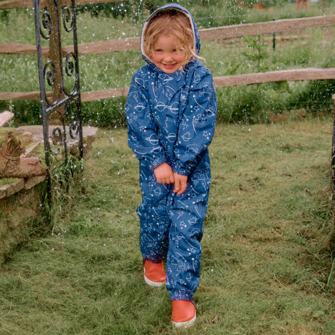 A child in a Navy 3 in 1 Waterproof Scampsuit stands on grass, smiling in the rain. The suit has a hood and a pattern of planets and stars.